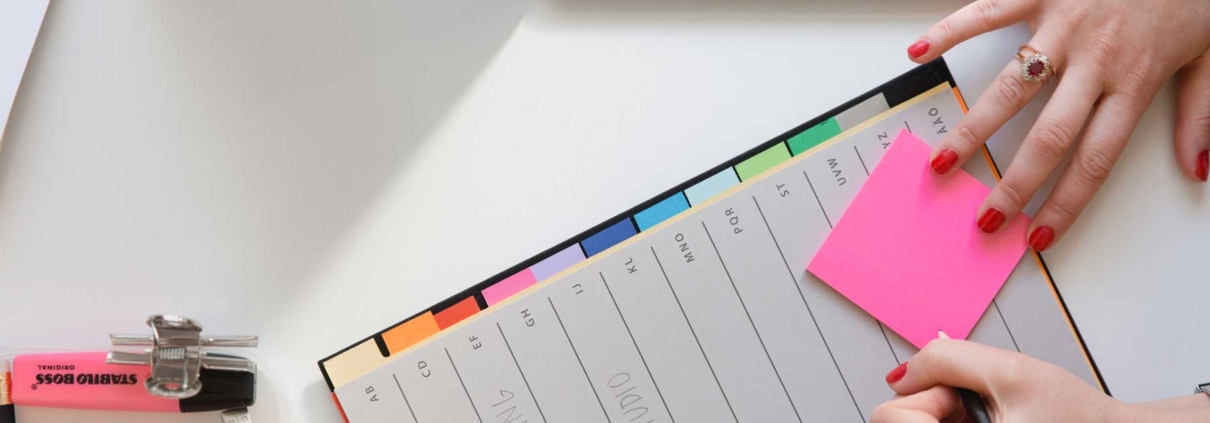A woman prepare to write on a pink post-it note while at her desk.